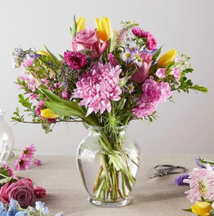 A vibrant and lush pastel flower bouquet in a clear glass vase featuring pink chrysanthemums, purple roses, and yellow tulips on a neutral tabletop.