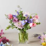 A lush pastel flower bouquet in a clear glass cylindrical vase, featuring pink tulips, purple irises, light pink roses, and lavender chrysanthemums accented with green foliage, set against a neutral grey background with florist shears and loose petals nearby.
