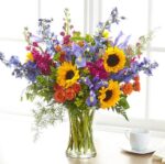 A vibrant and large floral arrangement in a clear glass vase featuring bright sunflowers, purple irises, blue delphiniums, orange spray roses, and magenta stock flowers, set against a bright window background.
