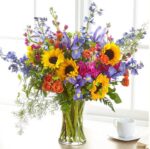 A vibrant and large floral arrangement in a clear glass vase featuring bright sunflowers, blue irises, orange spray roses, purple stock, and tall blue delphiniums with lush greenery.