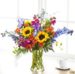 A vibrant and colorful mixed floral arrangement in a clear glass vase featuring sunflowers, orange roses, blue delphinium, purple irises, and magenta stock flowers, set against a bright window background.