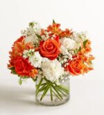 A vibrant floral arrangement featuring bright orange roses, orange alstroemeria, white carnations, and baby's breath in a clear glass cylinder vase.