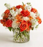 A lush "Sweet Citrus" floral arrangement in a clear glass cylinder vase, featuring vibrant orange roses, orange alstroemeria, white stocks, and delicate baby's breath.
