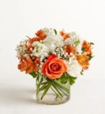 A vibrant floral arrangement featuring a prominent bright orange rose, surrounded by white carnations, orange alstroemeria, and delicate baby's breath in a clear glass cylinder vase.