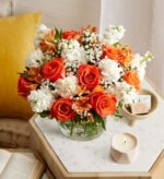A vibrant bouquet of orange roses, white carnations, and orange alstroemeria in a clear glass vase, placed on a marble side table next to a lit candle and an open book.