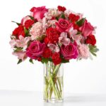 A lush bouquet of pink roses, red carnations, and light pink alstroemeria arranged in a clear cylindrical glass vase filled with water.