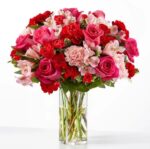 A lush and vibrant floral arrangement featuring a mix of pink roses, red carnations, and light pink alstroemeria in a clear cylindrical glass vase.