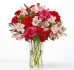 A vibrant floral arrangement in a clear cylindrical glass vase featuring pink roses, red carnations, and light pink alstroemeria (Peruvian lilies) with green foliage against a plain white background.