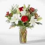 A festive floral arrangement in a clear glass cylinder vase featuring white lilies, red roses, red hypericum berries, and white stock flowers mixed with winter greenery like cedar and eucalyptus.