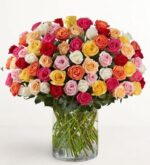 A large, dense bouquet of 100 multicolored premium long-stem roses arranged in a clear glass cylindrical vase against a white background.