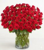 A large, dense bouquet of 100 premium long-stem red roses in a clear glass cylindrical vase against a plain white background.