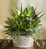 A lush memorial dish garden featuring a variety of green indoor plants in a white textured pot with a sentimental inscription.