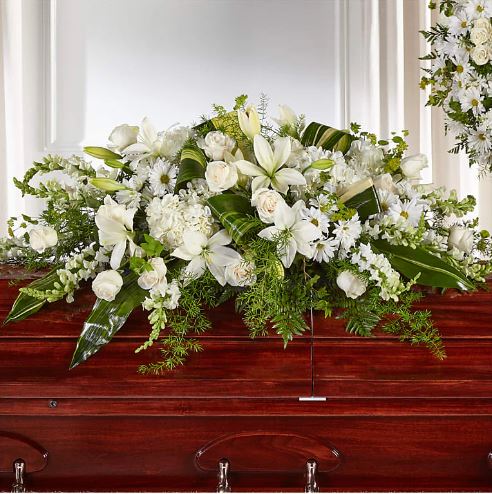 A premium, large horizontal casket spray composed of an abundance of white flowers including lilies, roses, carnations, and daisies, accented with lush green foliage and large variegated leaves, resting on a polished dark wood casket.