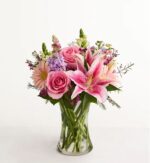 A vibrant "Always On My Mind" deluxe flower arrangement in a clear glass hourglass vase, featuring pink roses, Stargazer lilies, gerbera daisies, and purple stock.