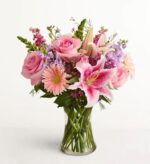 A premium floral arrangement in a clear glass vase featuring pink roses, pink lilies, peach gerbera daisies, and purple snapdragons against a plain white background.