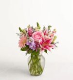 A lush floral arrangement in a clear glass vase featuring pink roses, stargazer lilies, pink gerbera daisies, and purple stock flowers against a plain white background.
