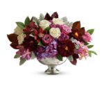 A luxurious harvest-themed floral arrangement in a silver pedestal bowl, featuring a rich palette of deep burgundy dahlias, cream roses, purple hydrangea, and pink orchids accented with dusty miller and dark autumn foliage.