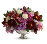 A lush and elegant floral centerpiece in a silver mercury glass pedestal bowl, featuring a rich palette of burgundy dahlias, purple hydrangeas, cream roses, and pink orchids accented with silvery dusty miller leaves.