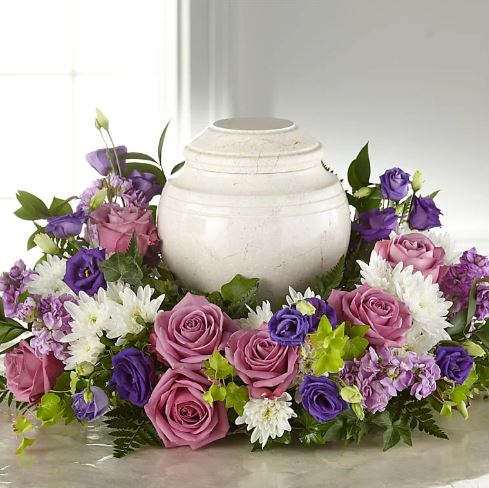A circular sympathy floral arrangement in shades of purple, lavender, and white surrounding a white marble cremation urn.