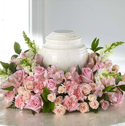 A lush circular floral arrangement featuring light pink roses, carnations, and alstroemeria surrounding a white marble cremation urn on a reflective surface.