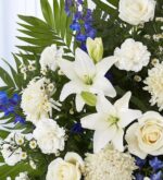 A lush funeral standing spray featuring white lilies, roses, carnations, and chrysanthemums accented with vibrant blue delphiniums and green palm leaves.