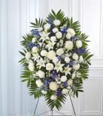 A large, elegant standing funeral spray arrangement featuring a mix of white lilies, roses, carnations, and chrysanthemums accented with vibrant blue delphinium and lush emerald green palm fronds on a wire easel.