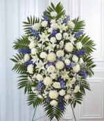 A large, symmetrical funeral standing spray arrangement featuring white lilies, roses, and chrysanthemums accented with vibrant blue delphinium and lush green palm fronds on a wire easel.