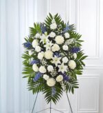 A tall, oval-shaped funeral standing spray arrangement featuring a mix of white lilies, roses, carnations, and chrysanthemums accented with vibrant blue delphinium and lush green palm fronds, displayed on a wire easel against a white paneled wall.