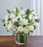 A large, lush bouquet of white roses, carnations, lilies, and snapdragons accented with green eucalyptus leaves in a glass cylinder vase lined with green leaf wrap.