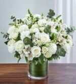 A lush and elegant bouquet of all-white flowers including roses, carnations, and alstroemeria, accented with fresh eucalyptus leaves in a glass vase lined with green leaf wrap.