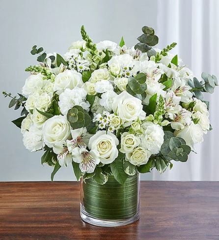 A lush and elegant bouquet of all-white flowers including roses, carnations, and alstroemeria, accented with fresh eucalyptus leaves in a glass vase lined with green leaf wrap.