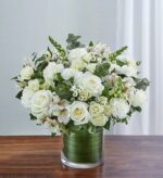 A lush and elegant bouquet of white flowers, including large roses, carnations, spray roses, and alstroemeria with purple accents, arranged in a glass cylinder vase lined with a green leaf-like texture.