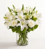 A premium floral arrangement featuring white lilies, roses, and snapdragons in a clear glass cylinder vase against a white background.