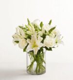 A deluxe classic all-white floral arrangement featuring white roses, lilies, and snapdragons in a clear glass cylinder vase against a minimalist white background.