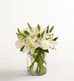 A sophisticated all-white floral arrangement featuring lilies, roses, and snapdragons in a clear cylindrical glass vase against a minimalist white background.