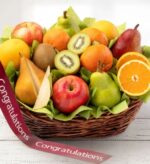 A premium wicker fruit gift basket filled with a variety of fresh orchard fruits including apples, pears, oranges, and sliced kiwis, adorned with a "Congratulations" ribbon.
