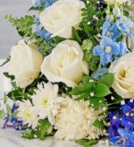 A close-up view of a serene funeral floral arrangement featuring large white roses, light blue delphiniums, white chrysanthemums, and lush green ferns against a soft grey background.