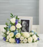 A serene blue and white floral cremation wreath or memorial arrangement featuring white roses, white chrysanthemums, and blue delphinium, surrounding a framed black-and-white portrait of an elderly man.