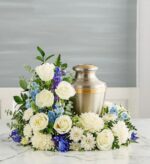 A serene blue and white floral cremation wreath featuring white roses, chrysanthemums, and blue delphinium surrounding a brushed metal urn with gold accents on a marble surface.