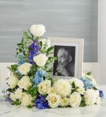 A premium blue and white floral cremation wreath or memorial arrangement featuring white roses, white chrysanthemums, and blue delphiniums surrounding a framed portrait of an elderly man.