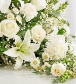 A lush, all-white crescent-shaped floral arrangement featuring lilies, roses, carnations, and baby's breath against a light background.