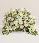 A large, crescent-shaped funeral floral arrangement featuring a dense mix of white lilies, roses, carnations, and snapdragons with lush green foliage.