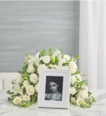 A crescent-shaped floral arrangement featuring all-white roses, lilies, and carnations surrounding a white portrait frame on a marble surface.