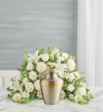 A crescent-shaped floral arrangement of white lilies, roses, and carnations surrounding a metallic cremation urn on a marble surface.