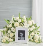 An elegant crescent-shaped funeral floral arrangement featuring a variety of premium all-white flowers, including roses, lilies, and carnations, surrounding a white-framed portrait of a woman.