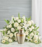 A lush, crescent-shaped memorial floral arrangement featuring all-white flowers like lilies, roses, and carnations surrounding a classic silver and gold cremation urn on a marble surface.
