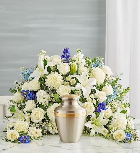 A crescent-shaped floral arrangement featuring white lilies, roses, and mums with blue delphinium accents, surrounding a silver and gold cremation urn on a marble surface.