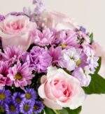 A close-up view of a lush "Daydream" flower bouquet featuring light pink roses, purple carnations, lavender daisies, and small blue asters with green foliage.