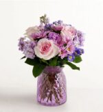 A lush floral arrangement featuring light pink roses, purple stock flowers, and lavender daisies in a textured purple glass vase.