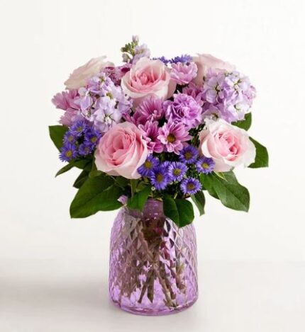 A lush floral arrangement featuring soft pink roses, purple chrysanthemums, and blue asters in a textured lavender glass vase.